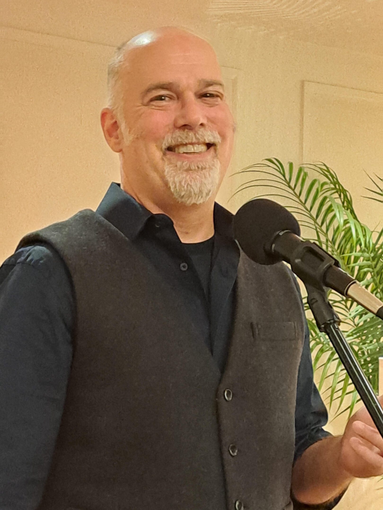 Smiling man speaking into a microphone with a plant in the background.