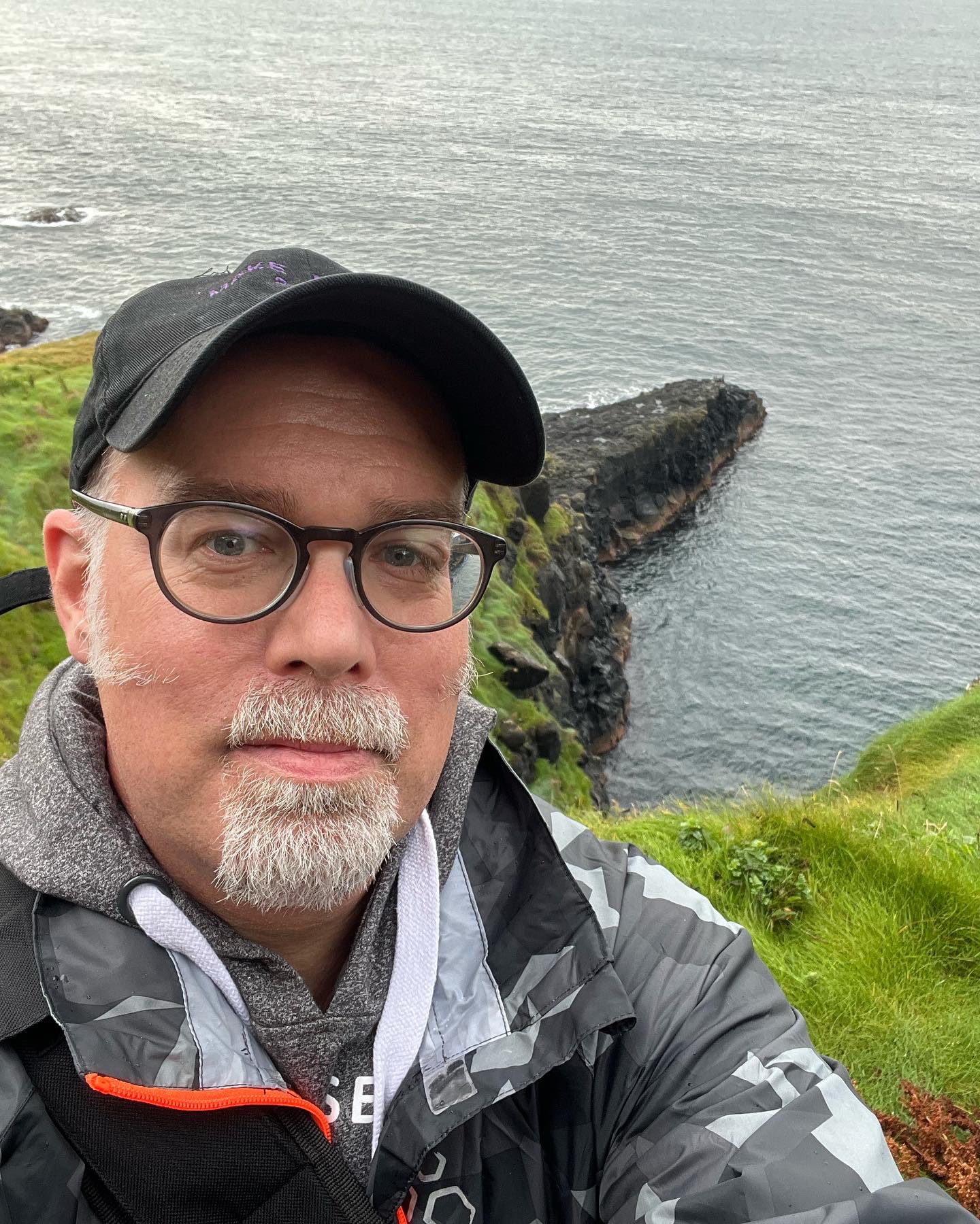 Man with glasses and cap standing on a grassy cliff by the water.